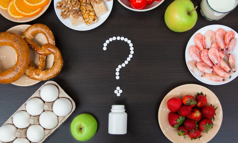 Alimentos comunes asociados a alergias e intolerancias (huevos, pan, frutilla, manzana, naranja, leche, frutos secos, mariscos) alrededor de un signo de pregunta de pastillas — Test de IgE específica (RAST), paneles IgG y estudios de celiaquía en Laboratorio Aclimu