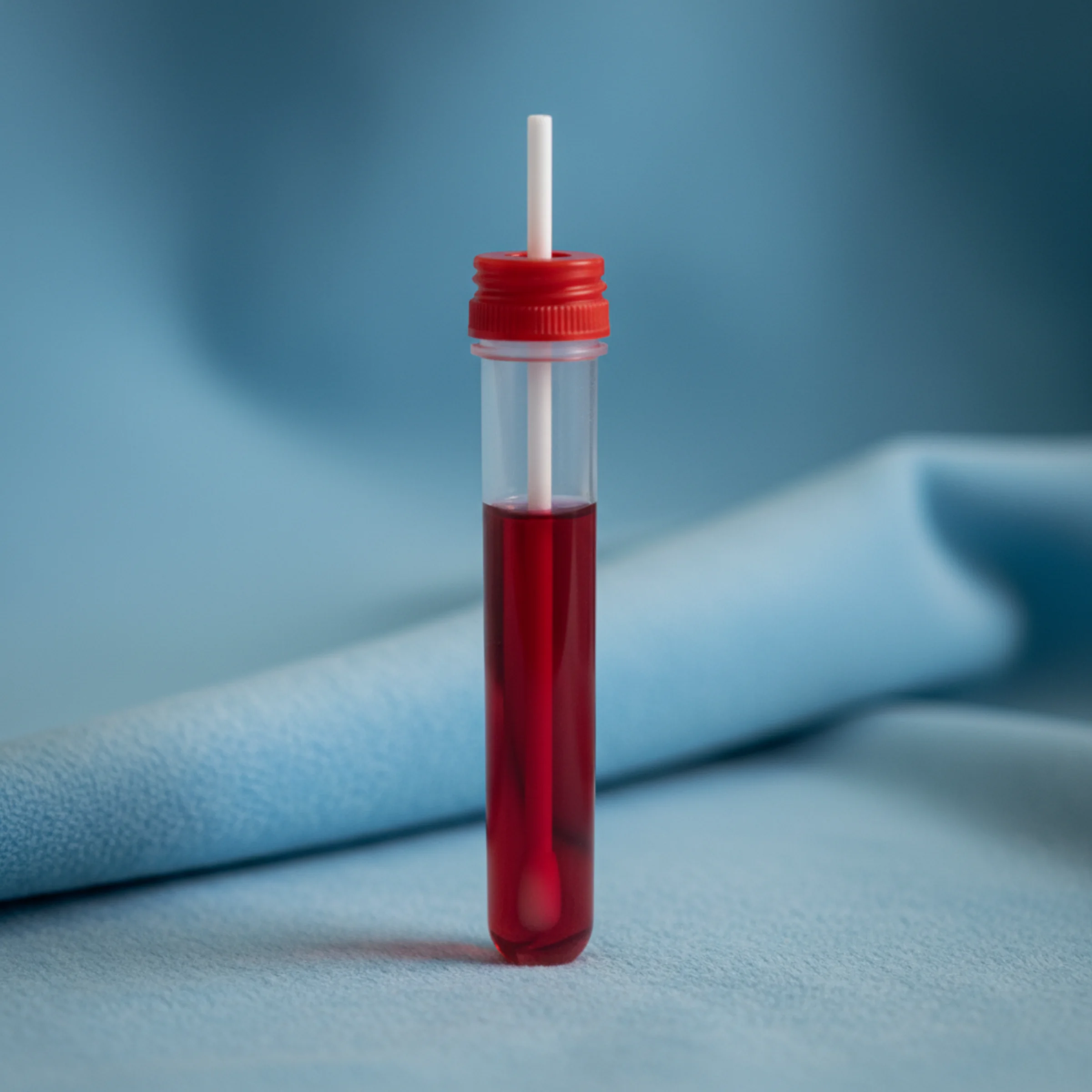 Hisopado COVID-19 por RT-PCR: tubo de transporte clínico con medio rojo VTM y hisopo estéril asomando por la tapa, sobre paño azul de laboratorio.