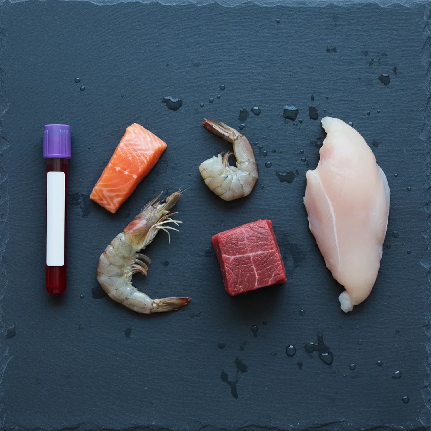 Panel 2 de intolerancia alimentaria IgG: tubo de extracción de sangre junto a salmón, camarones, carne vacuna y pollo sobre superficie de pizarra.